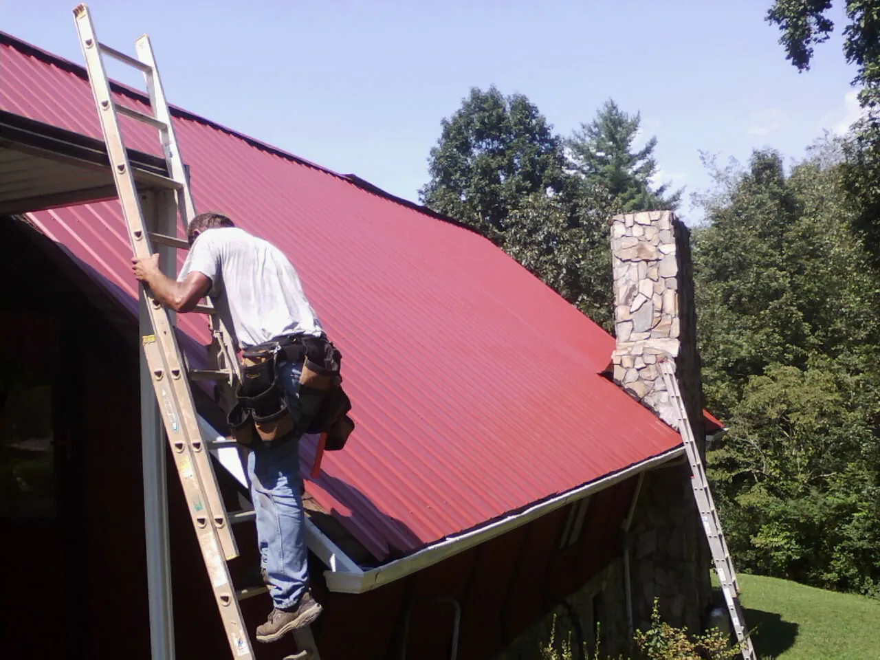 Roofer on a ladder beside a red metal roof and stone chimney — Soffit & Fascia Repair by Roofing Brothers in Belle Plaine